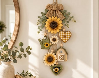Può includere: Una decorazione da parete sospesa con un grande girasole giallo, un annaffiatoio verde, un cuore a quadretti gialli, un piccolo fienile in legno e un cuore verde con un centro a quadretti bianchi e neri, arricchita da juta e fogliame.