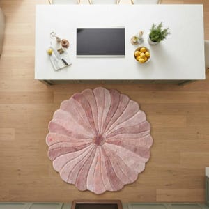 May include: A circular area rug featuring a scalloped, petal-like edge design in varying shades of soft pink. The rug has a central circular medallion with radiating tufted lines that extend outward to the rounded, overlapping petal borders.