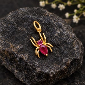 Puede incluir: Un colgante de araña de oro con una piedra preciosa rosa redonda para la cabeza y una piedra roja en forma de pera para el abdomen. La araña tiene ocho patas curvas y una pequeña anilla circular en la parte superior para sujetarla a una cadena.