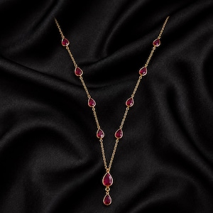 May include: A gold chain necklace featuring nine evenly spaced, teardrop-shaped red gemstones. The stones are set in gold bezels and arranged along the delicate chain, with a single dangling pendant at the center point of the necklace design.
