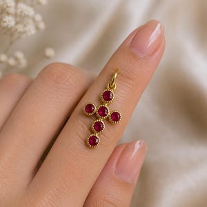 May include: A gold cross pendant featuring five round, deep red rubies set in a beaded metal frame. The pendant includes a small circular jump ring at the top for attachment, showcasing a detailed, textured gold border around each individual gemstone.