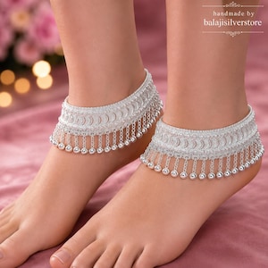 May include: A pair of silver anklets featuring a wide band with a repeating crescent moon pattern and a fringe of delicate silver chains ending in small, polished silver spheres. The text "handmade by balajisilverstore" is visible.