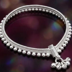 May include: A silver-toned anklet featuring a flexible snake chain base adorned with a continuous row of small, round beads along the edge. A cylindrical connector piece attaches a cluster of five small, spherical jingle bell charms.