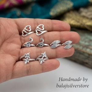 May include: A collection of six silver-toned metal rings featuring various designs including open heart shapes, detailed peacock motifs, textured leaf patterns, and floral arrangements with butterfly accents, all displayed together on a palm.