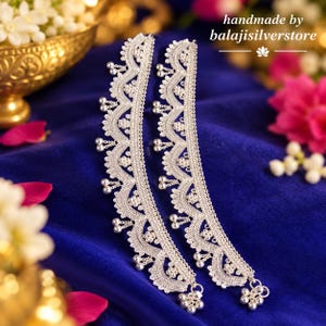 May include: A pair of silver anklets featuring an intricate scalloped lace-like pattern with small dangling spherical charms along the edges. The text "handmade by balajisilverstore" is visible in the upper right corner of the image.