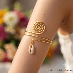 May include: A gold-toned metal arm cuff featuring a prominent spiral design at one end and a single, small, off-white cowrie shell charm dangling from the other end. The arm cuff is constructed from a smooth, continuous wire band.