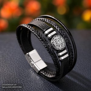 May include: A multi-strand black bracelet featuring braided leather cords and a smooth leather strip. The design includes a circular silver-toned charm with an intricate tree motif, flanked by two silver-toned metal accents and a magnetic clasp.