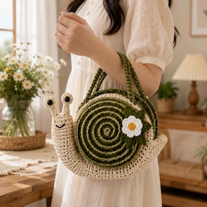 May include: A snail-shaped crochet handbag featuring a cream-colored body with two eye stalks and a dark green spiral shell. The shell is decorated with a white flower with a yellow center and green leaves. It includes two green crochet handles.