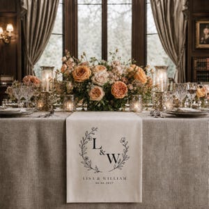 May include: A beige linen table runner featuring a black printed design of a floral wreath surrounding the letters L & W, with the text LISA & WILLIAM and the date 06.06.2027 printed below, displayed on a table with a floral centerpiece and candles.
