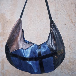 Puede incluir: Un bolso de hombro estilo hobo con un diseño de patchwork de paneles texturizados en azul metálico, bronce y negro. El bolso incluye una correa de hombro única y está acentuado con bandas negras lisas horizontales y verticales al frente.