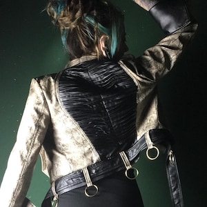 Peut inclure: Veste courte dorée et noire avec un dos en cuir noir et une ceinture à anneaux dorés. La veste a des manches longues avec des poignets évasés. La modèle porte une mini-jupe noire.