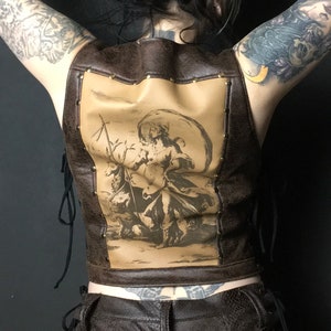 May include: A brown leather vest with a lace-up back and a vintage-style print of a woman on horseback. The vest has gold-colored metal accents.