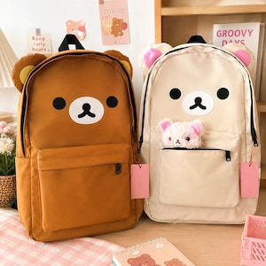 May include: Two backpacks featuring bear-themed designs. The left backpack is brown with yellow-accented ears, while the right one is cream-colored with pink-accented ears and a small plush bear toy tucked into the front zippered pocket.