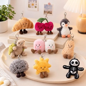 Può includere: Una collezione di nove portachiavi in peluche raffiguranti vari personaggi tra cui un croissant, ciliegie, un pinguino, una tartaruga, due marshmallow, un'arachide, una forma grigia testurizzata, un sole giallo e uno scheletro, con gancio.
