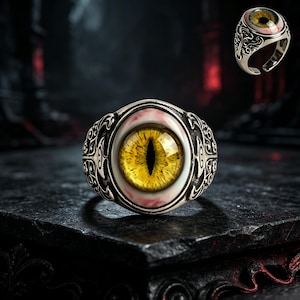 Può includere: Un anello in metallo color argento con un elemento centrale ovale che raffigura un realistico occhio di rettile giallo. La fascia presenta complessi motivi a volute in rilievo sui lati, che incorniciano l'occhio centrale con pupilla verticale.