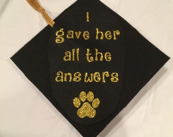 May include: Black graduation cap with gold glitter text that reads "I gave her all the answers." A gold glitter paw print is below the text. A gold tassel hangs from the cap.