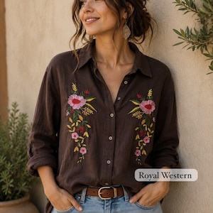 Puede incluir: Una camisa de manga larga con botones en color marrón oscuro que presenta un bordado floral colorido en la parte delantera. El bordado incluye flores rosas con hojas amarillas y verdes. La camisa se combina con vaqueros azul claro y un cinturón marrón.