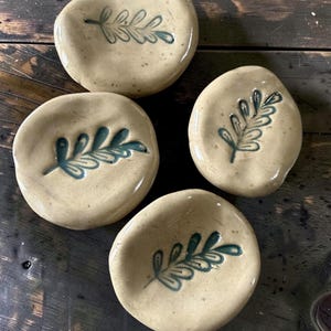 Peut inclure: Quatre petits plats en céramique beige, ronds, aux bords irréguliers et organiques. Chaque plat présente une empreinte de feuille en creux de couleur bleu sarcelle foncé au centre. La surface en céramique est lisse et brillante.