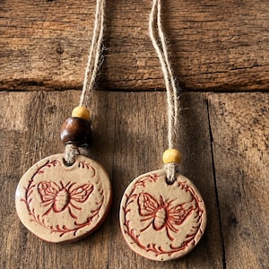 May include: Two circular clay ornaments featuring an embossed bee design surrounded by a leafy wreath. Each ornament is suspended by a natural jute twine string and accented with wooden beads in dark brown and light yellow tones.