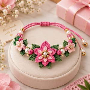 May include: A macrame bracelet featuring a central pink flower with gold and white beads, flanked by smaller pink flowers and green leaves. The bracelet is crafted from pink cord with adjustable ends finished with white pearls and gold beads.