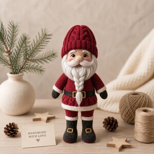 May include: A crocheted Santa Claus doll wearing a dark red cable-knit hat, a matching red sweater with a black belt featuring a gold buckle, and dark gray boots. The doll has a long white braided beard and a small card reading HANDMADE WITH LOVE.