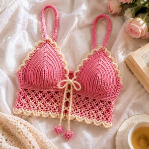 Pode incluir: Um top curto de crochê rosa com uma borda festonada na cor creme, um fecho frontal de amarrar com cordões creme e dois pequenos pingentes em forma de coração rosa nas pontas. A peça possui duas alças e uma base de crochê com padrão floral.