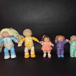 May include: A set of five small plastic dolls. The first has blonde yarn hair and a blue outfit. The second is bald with a yellow striped suit. The third wears a peach dress. The fourth has orange hair and a purple suit. The fifth wears a mint green suit.