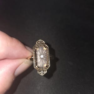 May include: A gold-toned ring featuring an elongated octagonal setting with a beaded border. The center contains a translucent, frosted stone with a sunburst pattern and a single small, clear, round-cut gemstone embedded in the middle.