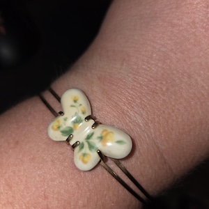 Könnte beinhalten: Ein dünner Armreif aus goldfarbenem Metall mit einem zentralen Anhänger in Schmetterlingsform. Der Anhänger besteht aus cremefarbenem Material mit einem bemalten Blumenmuster aus kleinen gelben Blüten und grünen Blättern auf beiden Flügeln.