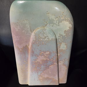 May include: A flat, rectangular ceramic vase with rounded top corners and a central arched relief. The surface features a mottled, weathered finish in soft pastel shades of mint green, pale pink, and cream with a textured, speckled appearance.