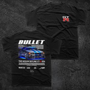 Può includere: Una t-shirt nera a maniche corte con un piccolo logo GT-R bianco e rosso sul petto. Il retro mostra una grande grafica di una Nissan Skyline GT-R R34 blu e argento con il testo BULLET e THE NISSAN SKYLINE GT-R R34 stampato sopra.