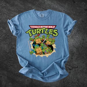 Könnte beinhalten: Ein hellblaues T-Shirt mit einer Grafik der vier Teenage Mutant Ninja Turtles-Figuren, die ihre typischen farbigen Masken und Waffen tragen. Der Text Teenage Mutant Ninja Turtles ist in großen grünen Buchstaben darüber gedruckt.