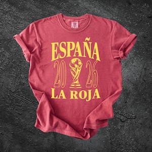 May include: A red short-sleeved t-shirt featuring yellow text that reads ESPAÑA 2026 LA ROJA with a central graphic of a trophy. The shirt has a crew neckline and rolled sleeves, displaying a weathered, vintage-style fabric texture throughout.