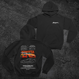 Peut inclure: Un sweat à capuche noir avec le logo Supra blanc sur la poitrine. Le dos présente un graphique technique de type plan d'une Toyota Supra MK4, incluant une illustration de voiture orange, des dessins au trait et le texte Toyota Supra MK4.