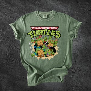 以下が含まれることがあります： セージグリーンの半袖Tシャツで、ティーンエイジ・ミュータント・ニンジャ・タートルズのグラフィックがプリントされています。4人のキャラクターがそれぞれの色のマスクと武器を身につけ、破れた紙のようなエフェクトと太字のテキストがデザインされています。