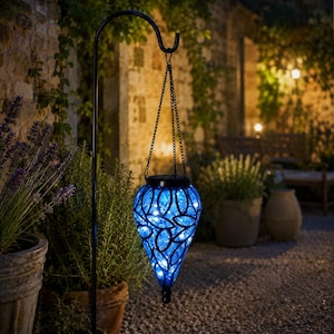 May include: A teardrop-shaped solar lantern with a blue glass body featuring a black metal cutout overlay. The lantern hangs from a black metal shepherd's hook by a thin chain and emits a soft blue light from internal string lights.