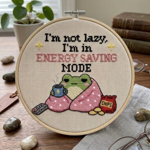 May include: A circular embroidery hoop containing a cross-stitch design on beige fabric. The text reads, "I'm not lazy, I'm in ENERGY SAVING MODE." A green frog wrapped in a pink polka-dot blanket holds a blue mug next to a bag of chips and a remote.