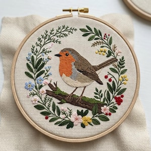 May include: A circular wooden embroidery hoop containing a cross-stitch design of a robin perched on a brown branch. The bird has an orange chest and brown wings. It is surrounded by a wreath of green leaves, small blue, pink, and yellow flowers.