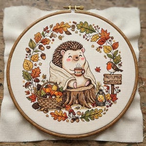 May include: A circular embroidery hoop containing a fabric piece with a stitched illustration of a hedgehog holding a mug labeled Cider, wrapped in a blanket on a tree stump, surrounded by a wreath of autumn leaves, a basket, and a sign reading Autumn Woods.