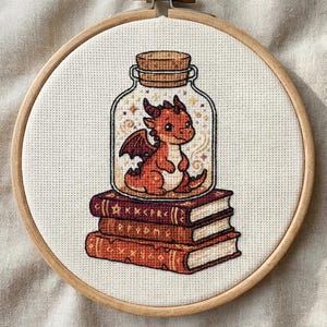 May include: A cross-stitch embroidery in a circular wooden hoop featuring a small orange dragon inside a glass jar, which is resting on a stack of three brown books with gold-colored runic symbols and star patterns on their spines and covers.