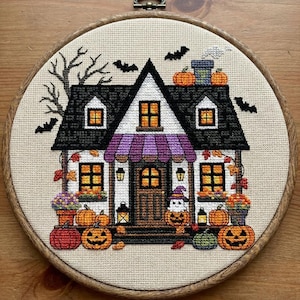 May include: A cross-stitch embroidery in a round wooden hoop depicting a two-storey house with a black roof, a purple striped awning, and glowing yellow windows. The scene includes orange pumpkins, a small ghost wearing a hat, and black bats.