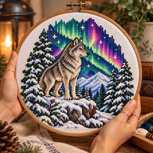 Puede incluir: Un bastidor de bordado circular de madera que contiene un patrón de punto de cruz de un lobo gris sobre un acantilado nevado. El fondo presenta una vibrante aurora boreal multicolor en el cielo nocturno sobre pinos y montañas nevadas.