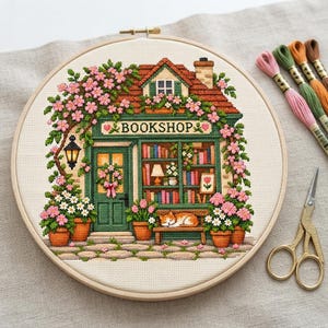 May include: A cross-stitch embroidery in a wooden hoop depicting a bookshop with a green storefront, a sleeping orange cat on a bench, and pink flowers. The word BOOKSHOP is stitched above the door. Embroidery floss and gold scissors are nearby.