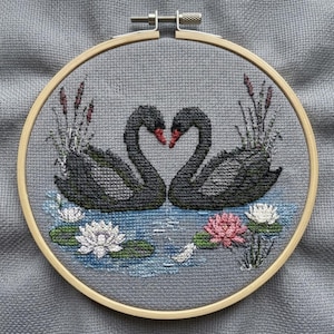 May include: A circular wooden embroidery hoop containing a cross-stitch design on grey fabric. The embroidery depicts two black swans facing each other, surrounded by white and pink water lilies, green lily pads, and tall brown cattails.