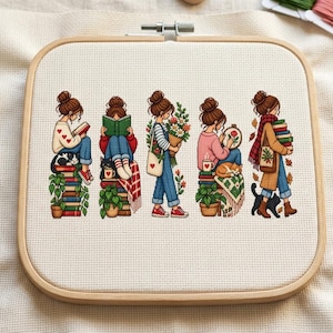 Può includere: Un telaio da ricamo quadrato in legno contenente un pezzo di tessuto Aida bianco con un motivo a punto croce. Il design presenta cinque illustrazioni di una persona con uno chignon castano, raffigurata mentre legge, tiene fiori, cuce e trasporta libri.