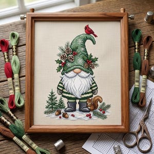 May include: A cross-stitch embroidery in a wooden frame depicting a gnome wearing a green hat decorated with pinecones, holly, and a red cardinal. The gnome has a white beard, striped sleeves, and grey boots, standing near a squirrel and pine trees.