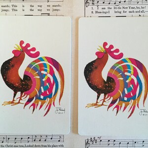 May include: Two playing cards with a colorful rooster illustration. The roosters have red, orange, and brown bodies with rainbow-colored tail feathers. The cards are rectangular with rounded corners and are set against a backdrop of sheet music.