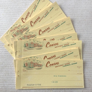 May include: A stack of vintage bank checks from Conroe National Bank, Conroe, Texas. The checks are a pale yellow with the bank's name in a stylized font and a building illustration. The checks are fanned out.