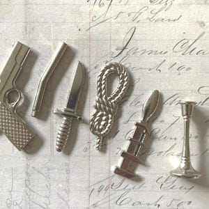 May include: A collection of small, silver-colored metal charms. The charms include a pistol, a knife, a rope knot, a spoon-like object, a candlestick, and a curved tube. The items are arranged on a vintage paper background.