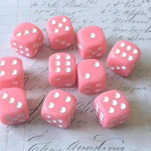May include: A collection of ten pink dice with white dots, arranged on a vintage paper background. The dice are a soft, pastel pink and the dots are clearly visible. The paper has handwritten text and numbers.